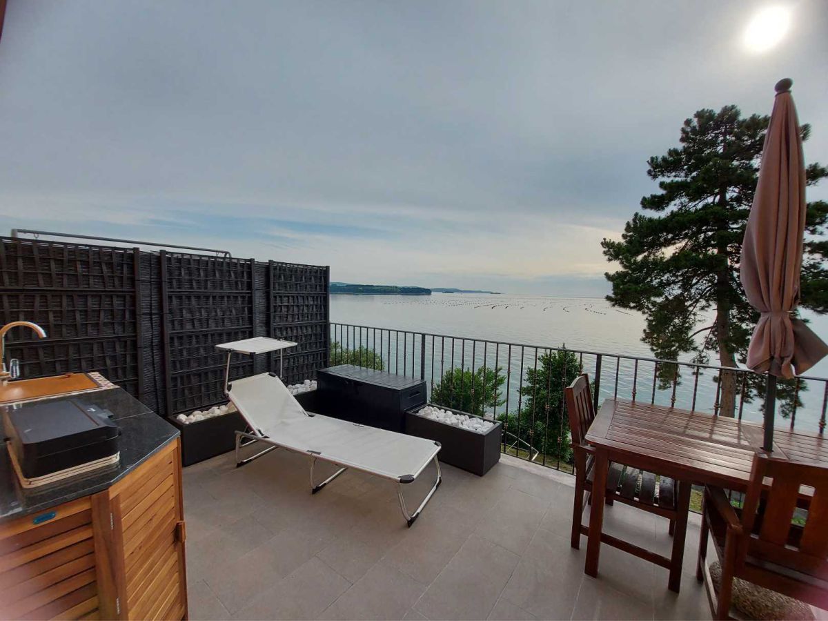 Terrace with kitchenette, deckchair, parasol, table with chairs, a tree with bushes, the sea in the background